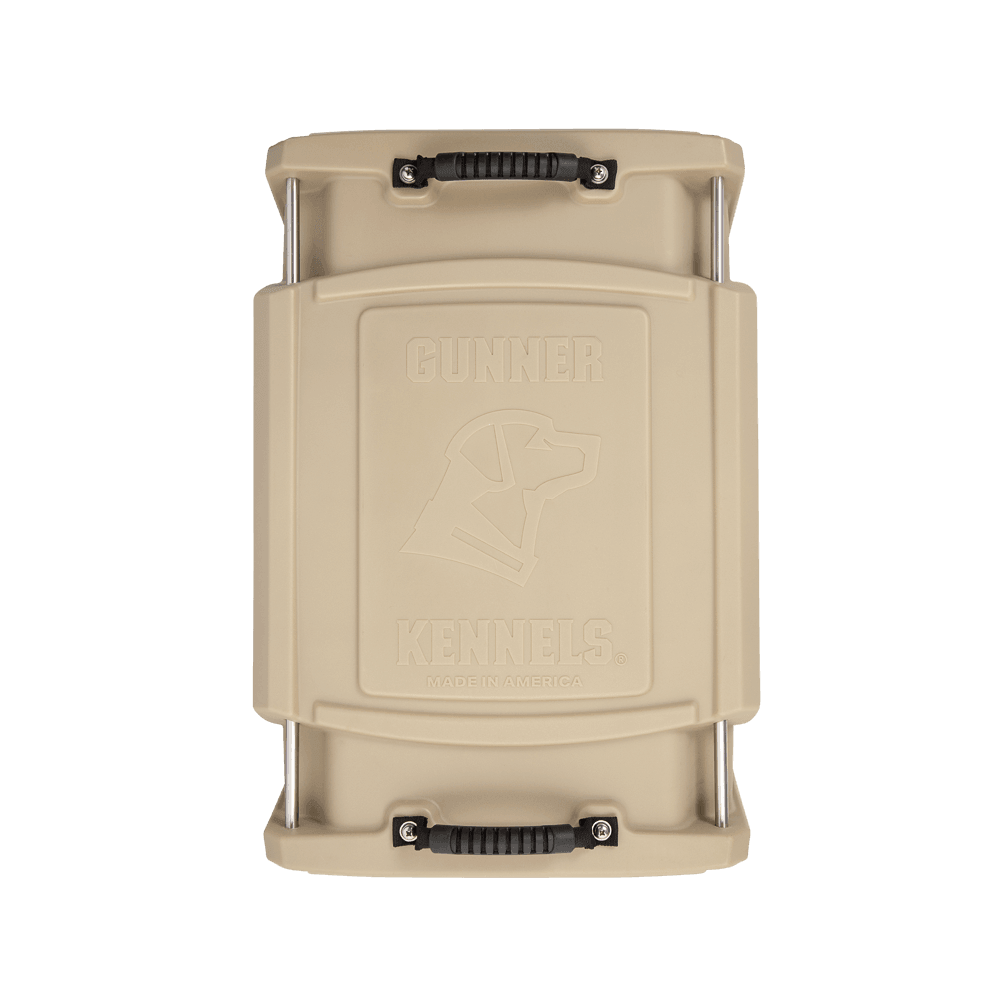 The GUNNER G1™ KENNEL - TAN is a beige, hard-sided dog crate with black handles at each end and an embossed dog head on the lid. Its double-walled design provides shock protection for enhanced safety.