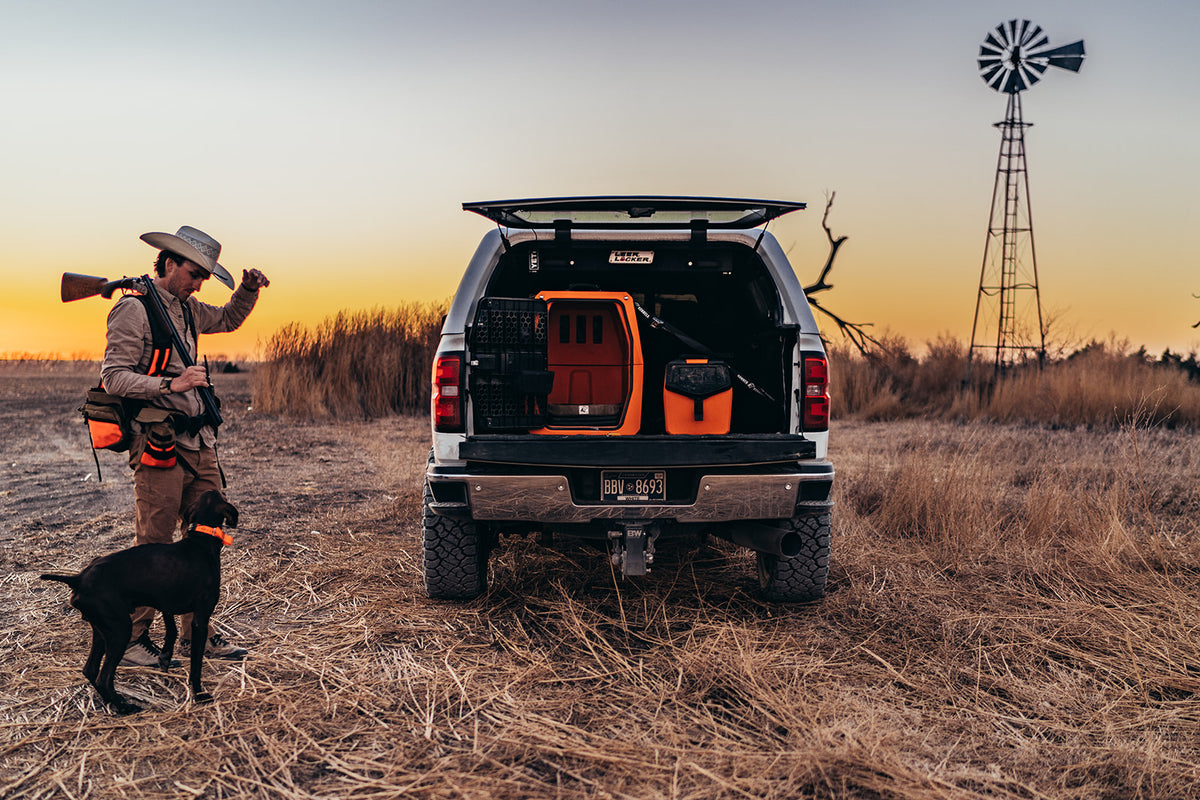A hunter with a rifle, orange vest, and cowboy hat stands beside a loyal hunting dog near a pickup truck loaded with outdoor gear, set in a dry field at sunset with a windmill in the background.