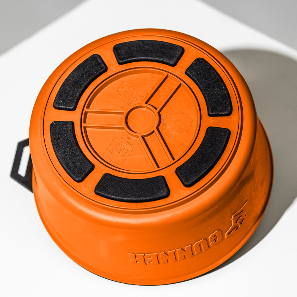 A GUNNER DOG BOWL - BLAZE ORANGE from GUNNER’s limited-edition Color Fetch collection is upside down on a white surface, showing black non-slip grips, embossed symbols on the base, and the GUNNER logo on the side.