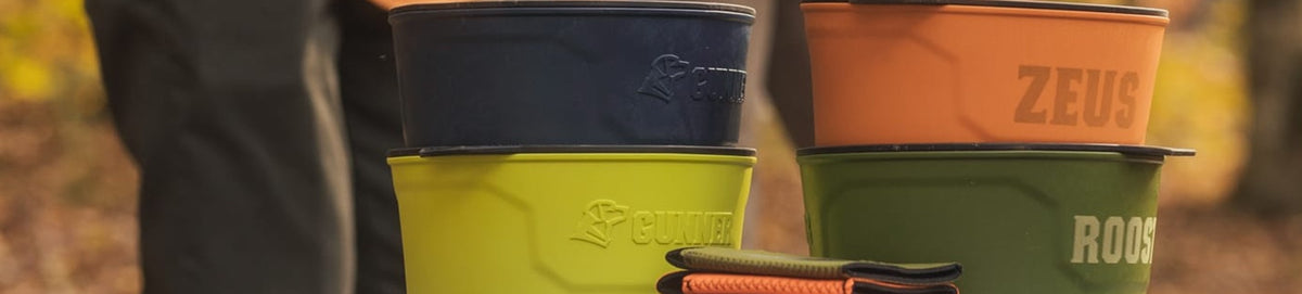 Four sturdy, colorful dog bowls labeled with names like ZEUS and ROCKET are stacked outdoors near a working dog crate, with a persons legs and a blurred forest background visible.