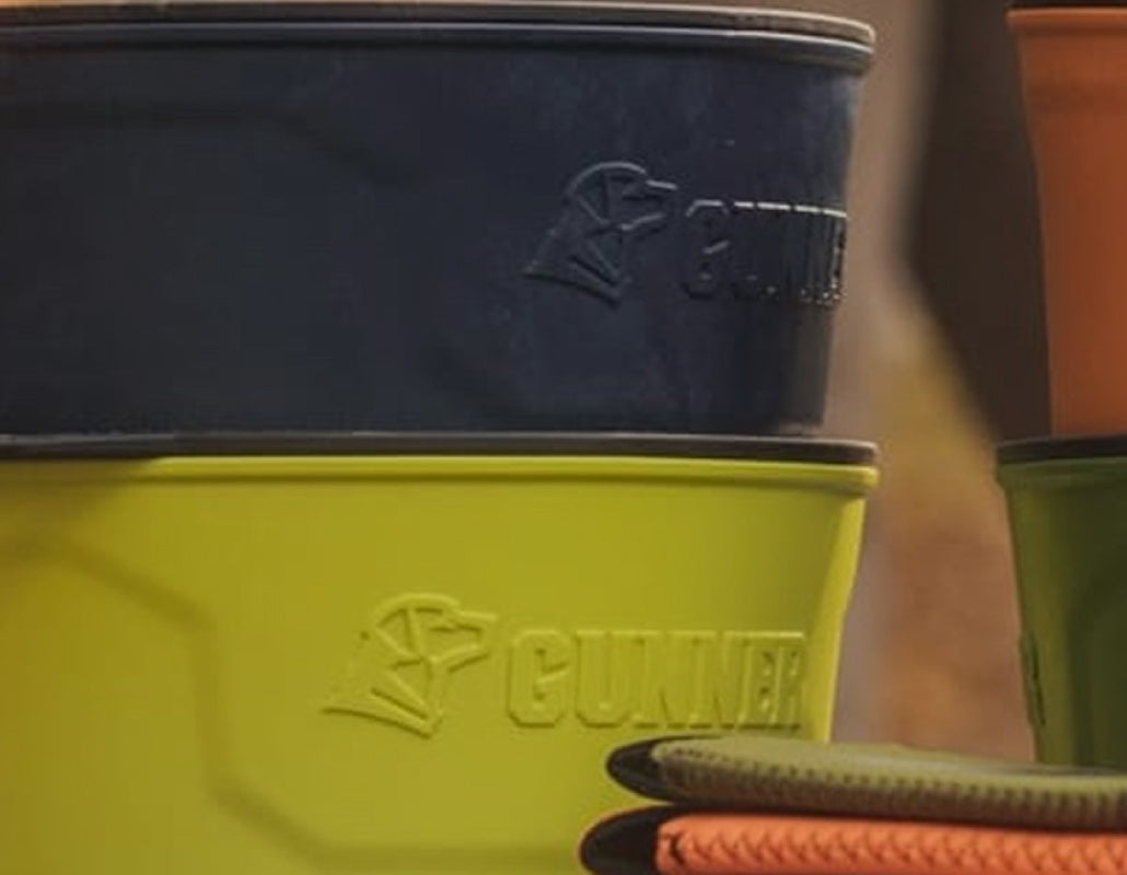 Close-up of stacked plastic bowls in navy blue, lime green, and orange, each embossed with a dog logo and the word GUNNER—perfect companions to your dog kennel or outdoor gear. The edge of a textured green item is partially visible on the right.