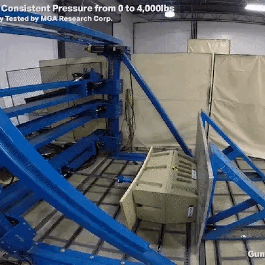 A large blue hydraulic press crushes a gun safe against a metal plate in a testing facility, demonstrating the safe’s strength and durability—ideal for securing outdoor gear or hunting dog supplies under heavy pressure.