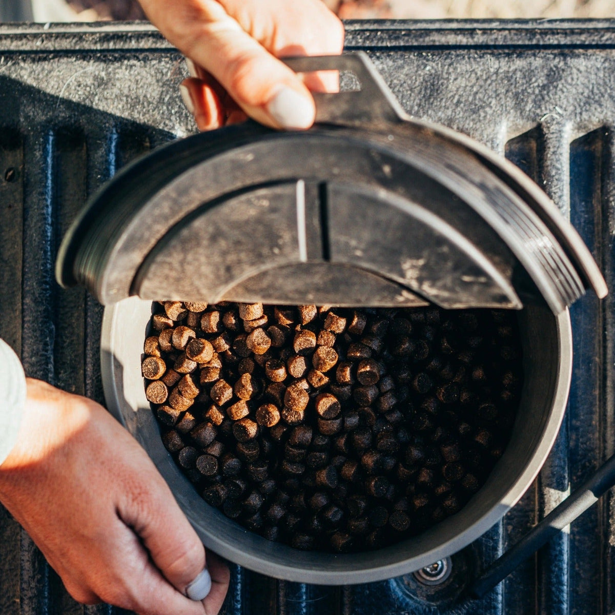 A person opens the GUNNER DOG BOWL - GUNMETAL by GUNNER, filled with dark brown, pellet-shaped pet food, on a textured dark surface.