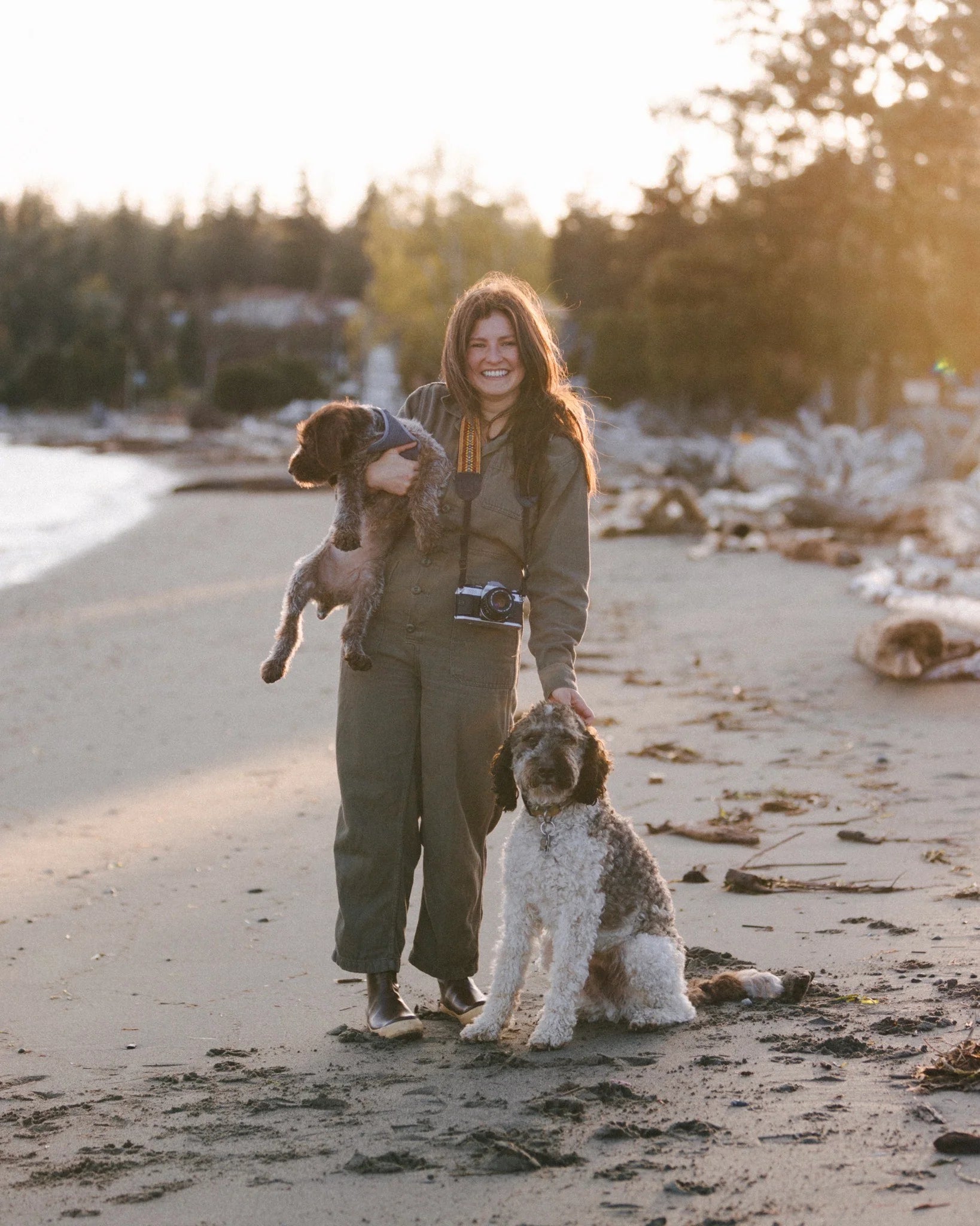 A woman stands on a sandy beach at sunset, smiling while holding one dog in her arms and touching another by her side. She wears outdoor gear and a camera around her neck, with trees in the background.