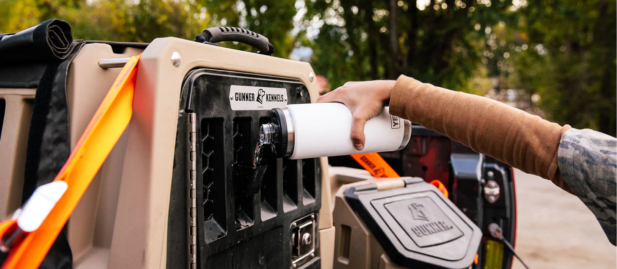 A person fills a white water bottle from a tap on the side of a rugged outdoor container attached to a vehicle, with trees, outdoor gear, and camping supplies in the background.