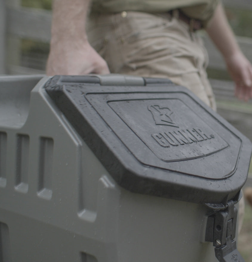 A person in tan pants carries a sturdy gray Gunner-branded dog kennel case with a black lid outdoors. Water droplets are visible on the surface, hinting at reliable protection for any hunting dog in rugged conditions.
