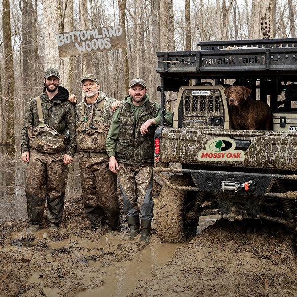Three men in camo gear stand in a muddy forest next to an off-road vehicle with a working dog riding in the back. A wooden sign reads Bottomland Woods. The truck and mens clothing are covered in mud.