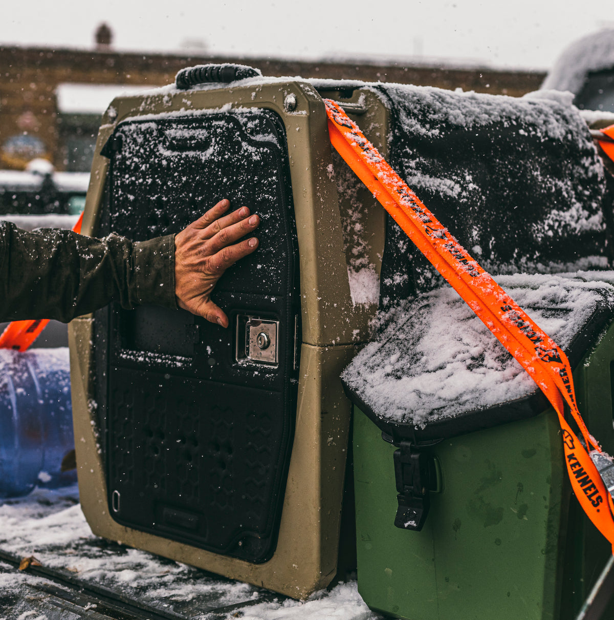 A hand touches a large GUNNER kennel secured with orange straps and dusted in snow outdoors, with other gear and the GUNNER COLD WEATHER DOOR COVER visible nearby.