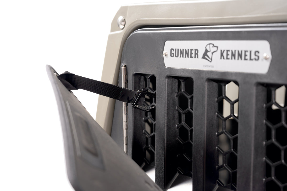 Close-up of a partially open GUNNER kennel door, showing its black metal grate, latch with strap, brand label, and the COLD WEATHER DOOR COVER made from thermoformed foam.