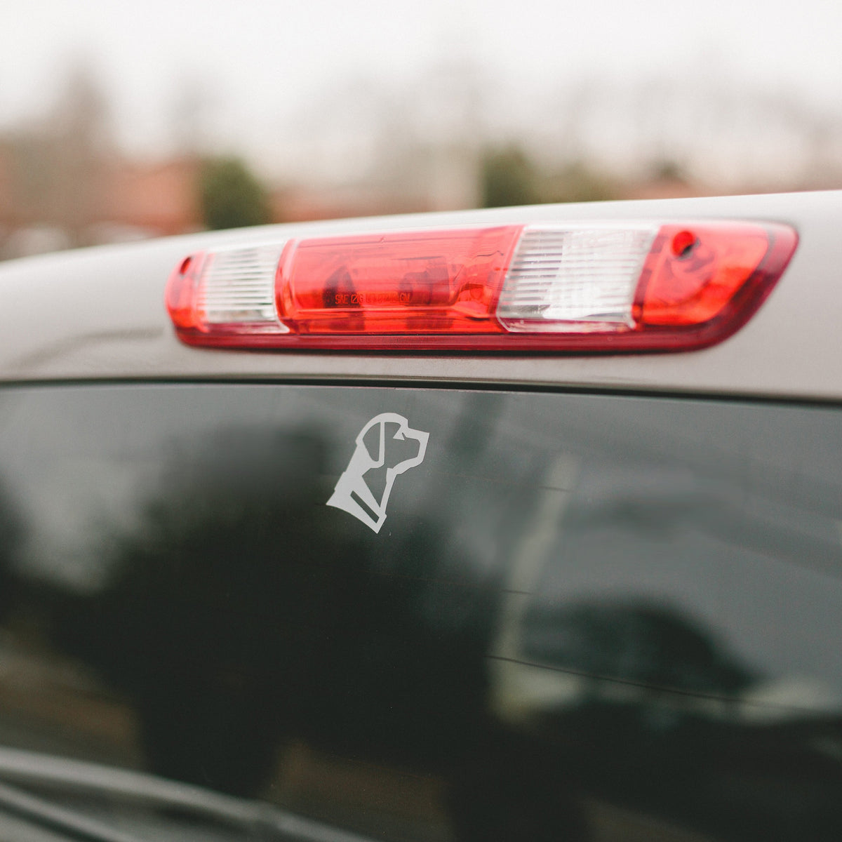 A GUNNER DOG HEAD DECAL, made of white vinyl and American-made, is shown on a cars rear window beneath the brake light, with blurred trees and buildings in the background.