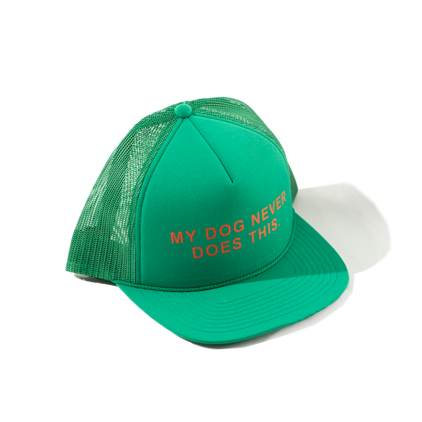 The GUNNER NEVER DOES THIS TRUCKER is a green mesh dog hat featuring MY DOG NEVER DOES THIS. in bold red capitals on the front, styled against a white background with a subtle shadow to showcase its playful look.